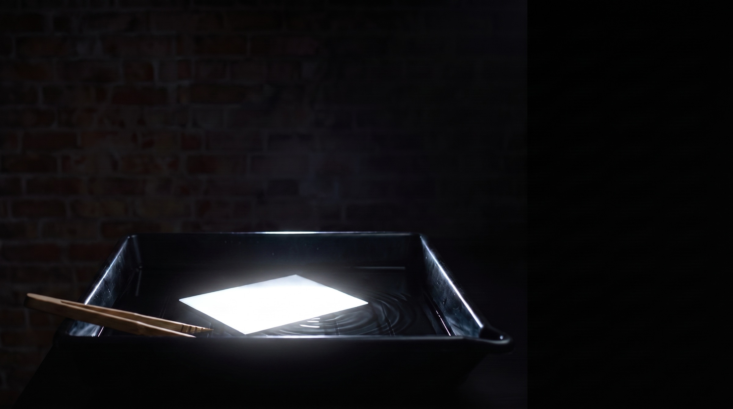 A glowing print floating in the developing bath, tongs resting on the tray